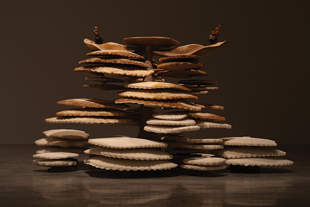 A sculptural installation featuring a layered structure of organic shapes. The sculpture is composed of stacked, textured elements in varying shades of brown and white, with colorful accents at the top.